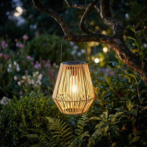 Lanterne solaire en rotin tressé Soléra suspendue à une branche dans un jardin la nuit, lueur chaude